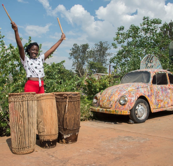 African American woman standing behind a set of African drums and a multi-colored car holding drumsticks in the air
