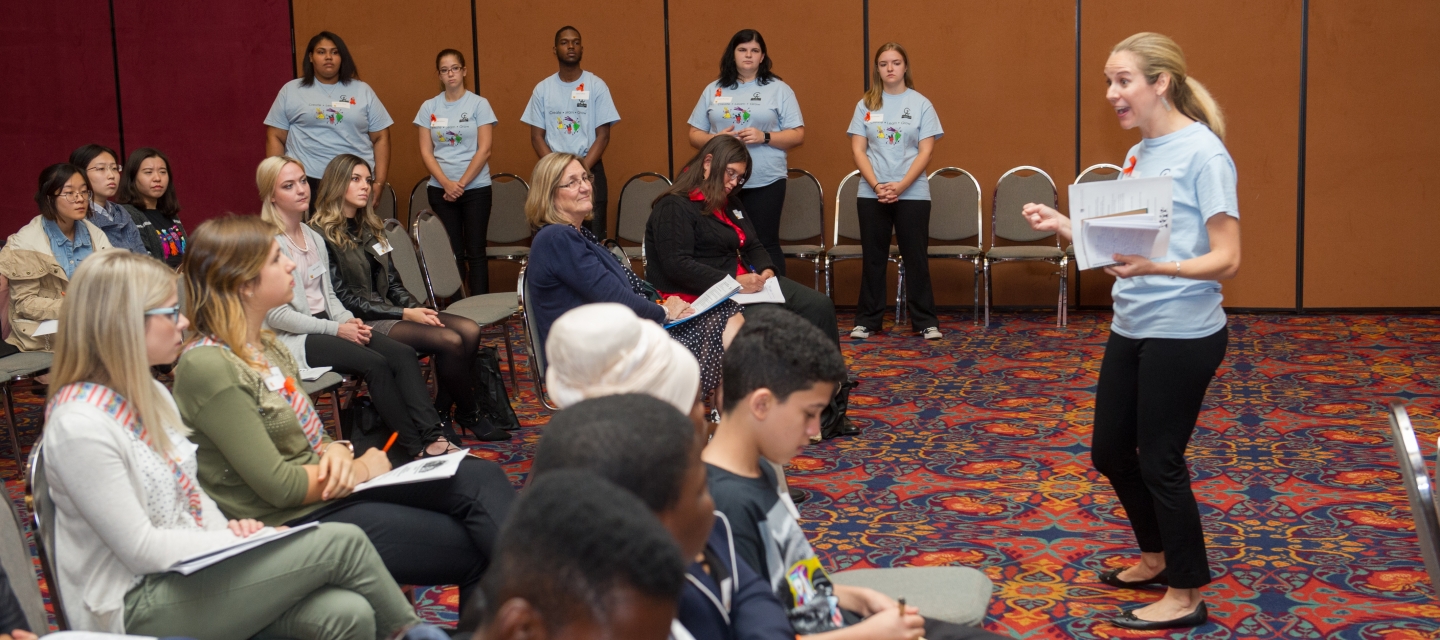 enthusiastic woman presenting to group of seated audience members while row of students stand near a wall