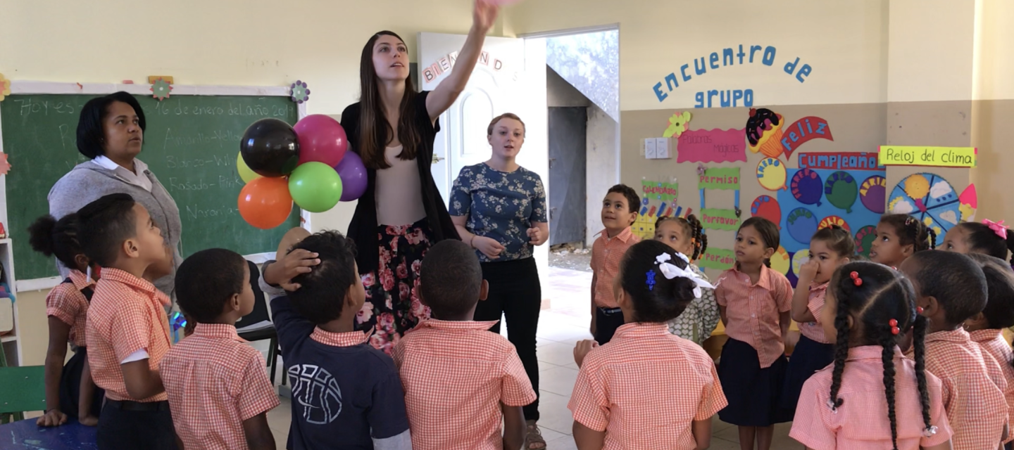 teacher tossing up pink balloon in middle of group of kids in orange uniforms